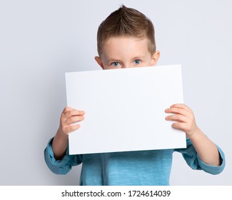 A Beautiful Boy Is Holding A Clean White Sheet Of Paper On An Isolated Background, Covering The Lower Half Of His Face With It. A Sly Look In A Child's Blue Eyes.