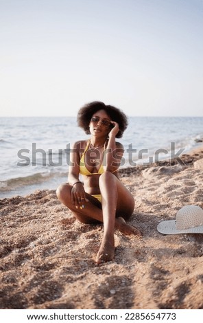 Beautiful brazilian adult young woman with afro curls and sunglasses in a yellow swimsuit enjoys the sea while sitting on the sand on a sea beach on a sunny day. Travel and relaxation, uniquesself