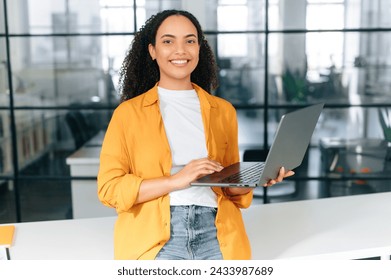 Beautiful brazilian or latino young professional business woman corporate marketing manager, company seo, holding portable computer and standing in a modern company office looking at camera, smile - Powered by Shutterstock