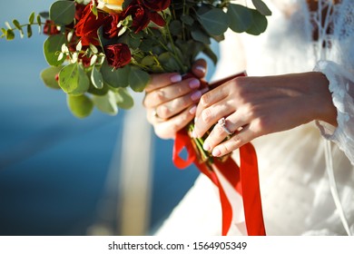 Beautiful bridal bouquet in bride's hands the background of sea and sun. Bride in an elegant white dress holds a stylish wedding bouquet with red and burgundy colors in her hands. Wedding day. 