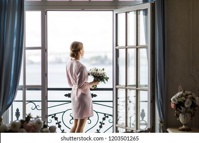 Beautiful bride in a bathrobe with a bouquet of roses is standing on the balcony. Morning of the bride. - Powered by Shutterstock