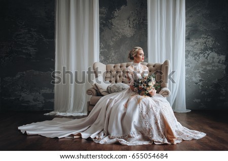 Beautiful bride in a cream wedding dress with a wedding bouquet, with orange and white flowers. Studio, gray background, modern, sofa, lace dress.
