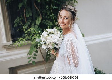 A Beautiful Bride With Curly Hair In A Lace White Dress With A Bouquet In Her Hands Stands Against The Background Of A Wall With A Growing Green Tree, A Bush On A Street Alley. Wedding Portrait.