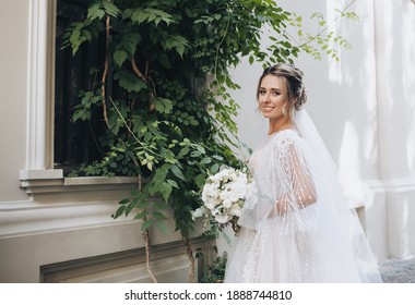 A Beautiful Bride With Curly Hair In A Lace White Dress With A Bouquet In Her Hands Stands Against The Background Of A Wall With A Growing Green Tree, A Bush On A Street Alley. Wedding Portrait.