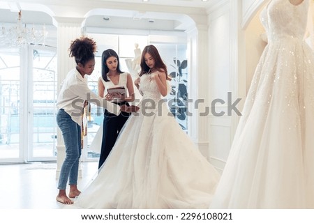 Beautiful bride discussing details with the wedding planner in studio. Attractive bridal shop owner women use digital tablet help customer choosing wedding gown at the store. Marriage ceremony concept