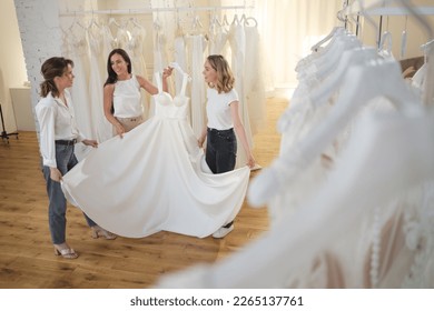 Beautiful bride getting dressed by her best friend in her wedding day and choosing a wedding dress in the shop and the shop assistant is helping her - Powered by Shutterstock