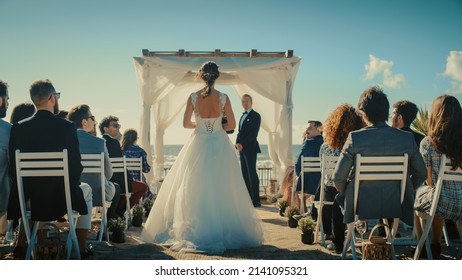Beautiful Bride in Gorgeous White Wedding Dress Going Down the Aisle, while Groom Waits at an Outdoors Ceremony Venue Near the Sea with Happy Multiethnic and Diverse Friends.