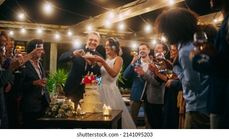 Beautiful Bride And Groom Celebrate Wedding At An Evening Reception Party With Multiethnic Friends. Married Couple Standing At A Dinner Table, Kiss And Cut Wedding Cake.