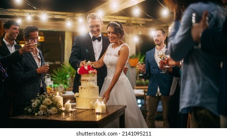 Beautiful Bride and Groom Celebrate Wedding at an Evening Reception Party with Multiethnic Friends. Married Couple Standing at a Dinner Table, Cutting Wedding Cake. - Shutterstock ID 2141095211