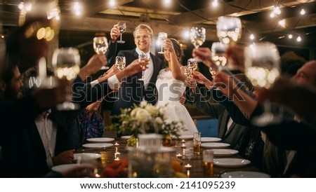 Beautiful Bride and Groom Celebrate Wedding at an Evening Reception Party. Newlyweds Propose a Toast to Happy Marriage, Standing at a Dinner Table with Best Multiethnic Diverse Friends.