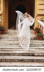 Beautiful Bride Having Fun On The Stairs Outdoor While Her Bridal Long Veil Flying On Wind In Air.