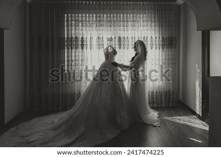 A beautiful bride with a long veil in her room, wearing a robe. Wedding dress on a mannequin. The bride in the morning before the wedding ceremony.