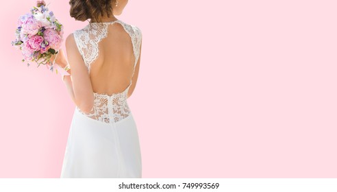 Beautiful Bride On A Pink Background. Woman Dressed In Wedding Dress With Lace And An Open Back. Bouquet In Hands.