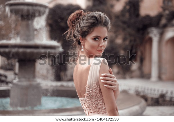 Beautiful bride with\
pearl earrings jewelry wears pink prom dress. Outdoor romantic\
portrait  of Attractive brunette woman with makeup and wedding \
hair style posing at\
park.