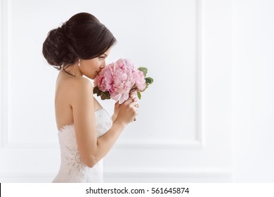 Beautiful bride perfect style. Wedding hairstyle make-up luxury wedding dress and bride's bouquet. Young attractive multi-racial Asian Caucasian model like a bride against white room smells at bouquet - Powered by Shutterstock