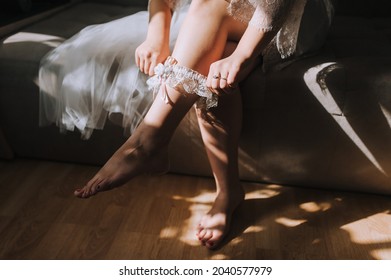 A beautiful bride puts a garter on her leg, sitting on the bed in the morning in the interior, room. Wedding photography.