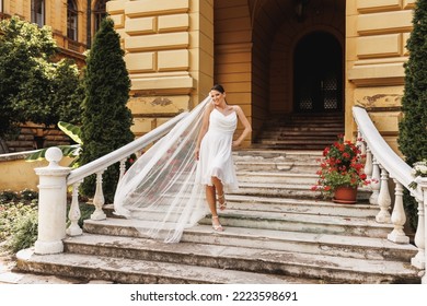 Beautiful Bride Walking Down The Stairs Outdoor While Her Bridal Long Veil Flying On Wind In Air.