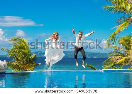 Beautiful bride in wedding dress with long train and groom jumping in the infinity pool in the hotel on a tropical island. Wedding and honeymoon.