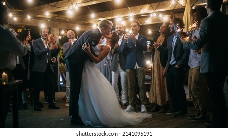 Beautiful Bride in White Dress and Groom in Stylish Black Suit Celebrate Wedding at an Evening Reception Party. Newlyweds Dancing at a Venue with Best Multiethnic Diverse Friends.