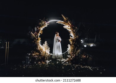 A Beautiful Bride In A White Long Dress Stands At Night Near A Luminous Round Arch Made Of Reeds Decorated With Lamps And Garlands. Wedding Photography, Portrait.