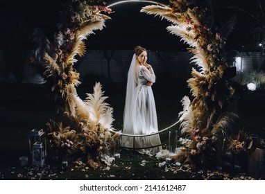 A Beautiful Bride In A White Long Dress Stands At Night Near A Luminous Round Arch Made Of Reeds Decorated With Lamps And Garlands. Wedding Photography, Portrait.