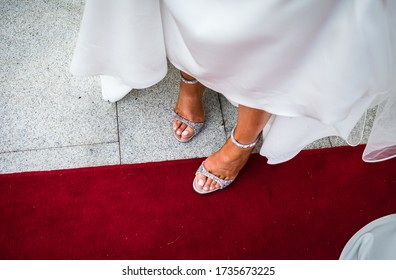 Beautiful Bride's Feet With White Nail Polish On The Toes, New Fancy Shiny High Heels Silver Shoes, Long White Silk Wedding Dress, Stepping On The Red Carpet