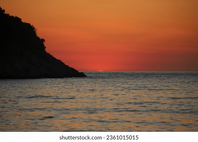 A beautiful bright and burnt Orange, Golden and Red Dusk Sunset over the Sea at Kakoma Bay in Albania with a Hillside in Black Silhouette and Clouds in the evening Sky. Adlı Stok Fotoğraf