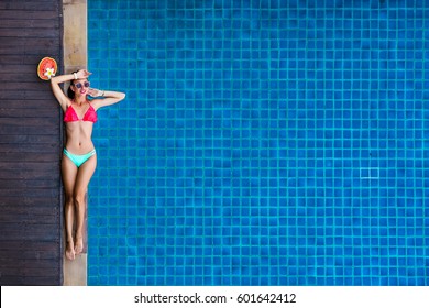 Beautiful Bright Sexy Funny Woman Lies On The Edge Of The Pool, Sunbathing, Enjoys. Perfect Body And Healthy Skin And Hair, Fashionable Bikini, Sunglasses, Watermelon. View From Above