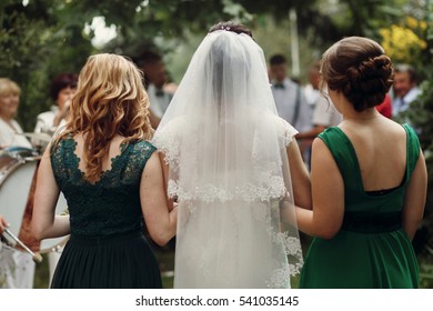 Beautiful Brunette Bride Under Veil In White Wedding Dress Walking With Two Gorgeous Bridesmaids In Green Dresses To Wedding Aisle, Shot From Behind