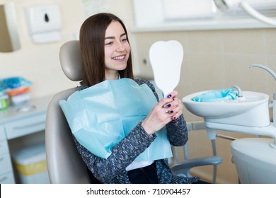 Beautiful Brunette Caucasian Girl In Dentist Blue Bib Sit In The Dentist Chair With Mirror In Hands, Looking At Herself, Smiling