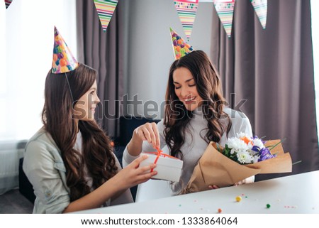 Beautiful brunette caucasian mother and daughter together in room. Female adult open present box and smile. She hold flowers. Her daughter give present to mom and smile.