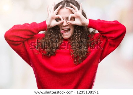 Beautiful brunette curly hair young girl wearing glasses and winter sweater over isolated background doing ok gesture like binoculars sticking tongue out, eyes looking through fingers. Crazy