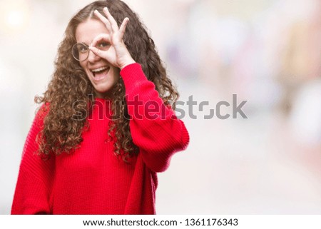 Beautiful brunette curly hair young girl wearing glasses and winter sweater over isolated background doing ok gesture with hand smiling, eye looking through fingers with happy face.