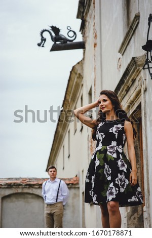 Beautiful brunette in a dress with flowers goes in the yard of an old castle. Stylish girl in a black dress on a background of gray vintage building