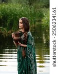 Beautiful  brunette extra long braided hair girl in Indian traditional dress posing near river with jug . Portrait of young attractive stylish woman in sari on  green nature background.