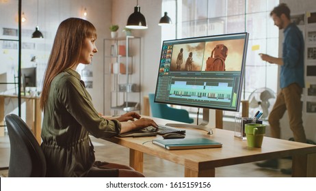 Beautiful Brunette Female Video Editor Works with Footage on Her Personal Computer with Big Display. She Works in a Cool Office Loft. Other Male Creative Colleague Walks in the Background.