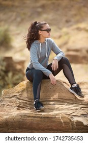 A beautiful brunette fitness model in black tights and a grey sports hoodie sits on a rock on a bright Sunny day in a desert area wearing sunglasses