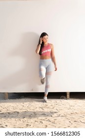 Beautiful Brunette Fitness Model And Yogi Wearing A Pink Crop Top And Gray Yoga Tights Standing Against A Plain Beige Wall