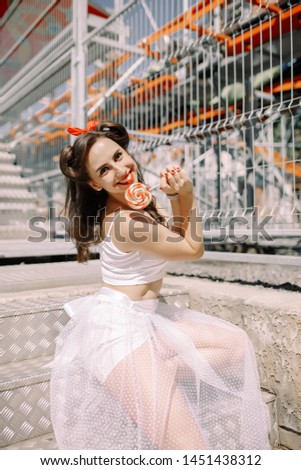 beautiful brunette girl in an amusement park in pin-up style with a coffee shop on the background of carousels with retro hairstyle in a white skirt. 
emotions. eat candy at an amusement park.