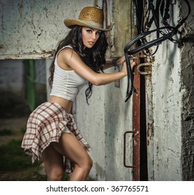 Beautiful brunette girl with country look, indoors shot in stable, rustic style. Attractive woman with cowboy hat, denim shorts and tight white top. American country style farmer near barn harness