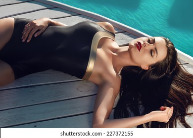 Beautiful Brunette Girl In Good Shape With Long Dark Hair And Tan Skin Red Lips In Black Swimming Suite With Golden Line With Modern Rings Lie Near The Pool With Green Water Closing Her Eyes And Smile