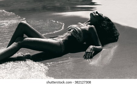 Beautiful Brunette Girl In Good Shape With Long Dark Hair And Tan Skin Red Lips In Black Swimming Suite With Golden Line With Modern Rings Lie On Beach Sand,with Green Water Closing Her Eyes And Smile