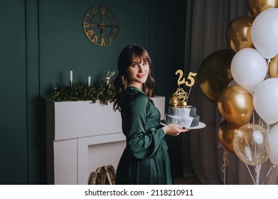 A beautiful brunette girl holds a delicious cake with the numbers 25 on her birthday on the background of the interior. Festive photo.