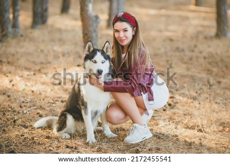a beautiful brunette girl in a short skirt and a burgundy leather jacket with a hoop on her head hugs a husky dog in the forest