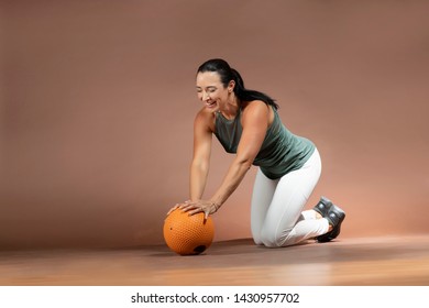 Beautiful Brunette Hair Fitness Model Wearing White And Green Sportswear  Doing A Knee Pushup On An Orange Medicine Ball On The Floor Against A Brown Background   