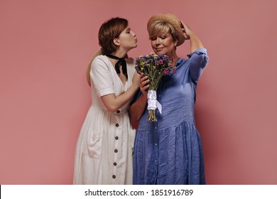 Beautiful Brunette Lady In White Clothes Blowing Kiss And Posing With Old Woman In Blue Dress And Hat With Wildflowers On Isolated Backdrop..