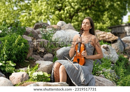 beautiful brunette in a linen dress with a violin on a pile of boulders at an old country house