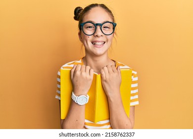 Beautiful brunette little girl wearing glasses and holding book smiling and laughing hard out loud because funny crazy joke. 
