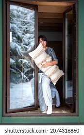 A Beautiful Brunette Middle-aged Woman In Glasses Standing In The Window Frame With A Woolen Cushion In Her Hands And Snowy Forest Reflected In A Window Glass 