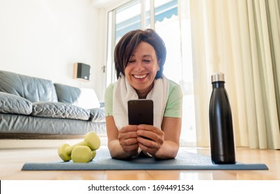 Beautiful brunette older woman training gym at home holding smartphone. Senior female workout with fitness app on the phone. Concept about people, sport, quarantine.